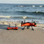 SwellPro Fisherman FD3 Waterproof Fishing Drone with 4K Camera sits on sandy beach terrain, accompanied by its red handheld radio controller, waves visible in the background.